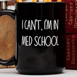 May include: Black ceramic mug with the text "I CAN'T, I'M IN MED SCHOOL" in white. The mug has a large handle and is next to two red books. Pencils are visible inside the mug.