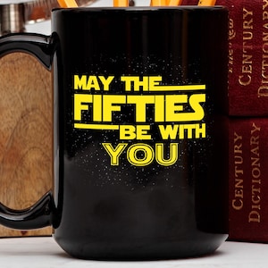 May include: Black ceramic mug with the text "MAY THE FIFTIES BE WITH YOU" in yellow, inspired by a popular science fiction franchise. The mug has a large black handle and is set against a wooden surface and two red books.