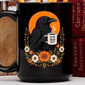 Puede incluir: Taza de cerámica negra con una ilustración de un cuervo sosteniendo una taza de café. La taza dice "IS HE DEAD YET?". El cuervo está sobre un círculo naranja, rodeado de elementos florales.