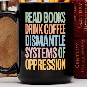 Könnte beinhalten: Schwarze Kaffeetasse mit dem Text "READ BOOKS DRINK COFFEE DISMANTLE SYSTEMS OF OPPRESSION" in bunten Blockbuchstaben. Die Tasse steht neben zwei roten Büchern mit goldener Schrift.