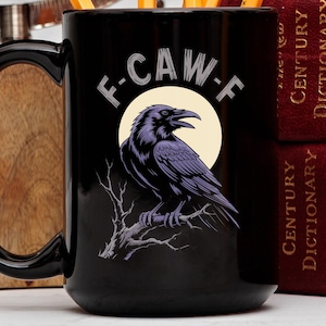 May include: Black ceramic mug featuring a purple raven perched on a branch against a yellow moon. The mug has the text "F-CAW-F" above the raven. A perfect gift for bird lovers.