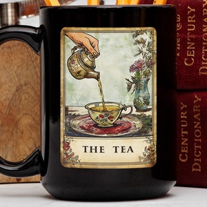 Peut inclure: Une tasse en céramique noire avec une illustration vintage d'une main versant du thé d'une théière dans une tasse à thé. L'illustration est encadrée d'une bordure dorée et le texte "THE TEA" est imprimé sous l'image.