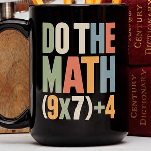 Puede incluir: Taza de cerámica negra con el texto "DO THE MATH" en un esquema de colores retro. La ecuación (9x7)+4 está debajo del texto. La taza tiene un asa grande y está junto a una superficie de madera y dos libros rojos.