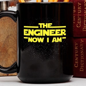 May include: Black coffee mug with a yellow graphic that reads "The Engineer - Now I Am -" against a black starry background.