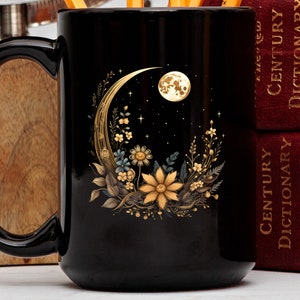 May include: Black ceramic mug with a crescent moon and floral design in gold. The moon has a face and is surrounded by stars. The mug has a handle and the words "21st Century Dictionary" are visible on the books behind it.