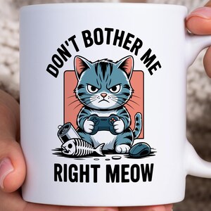 May include: White ceramic mug featuring a grumpy cartoon cat playing a video game, with the text "DON'T BOTHER ME RIGHT MEOW." The cat is blue and gray, holding a game controller. A fish skeleton and a mouse are also shown.