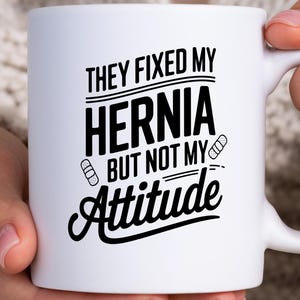 May include: White ceramic mug with the text "THEY FIXED MY HERNIA BUT NOT MY Attitude" in black. The design includes bandage graphics. The mug is held by a person.