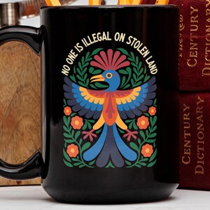 Könnte beinhalten: Schwarze Keramik-Tasse mit einem farbenfrohen Vogeldesign und dem Text "NO ONE IS ILLEGAL ON STOLEN LAND". Der Vogel hat blaue, orange und gelbe Federn, umgeben von grünen Blättern und orangefarbenen Blumen. Ein tolles Geschenk.
