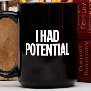 May include: Black ceramic mug with the white text "I HAD POTENTIAL". The mug has a large handle and is next to a stack of red books. Pencils are visible inside the mug.