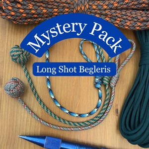 May include: Three begleri skill toys with monkey fist knots and paracord strings in orange, black, blue, and grey patterns. The text Mystery Pack Long Shot Begleris is overlaid. A blue metal pen and a coil of dark green paracord are also visible.
