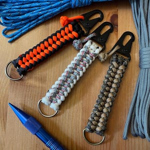 May include: Three paracord keychains with different color combinations. The keychains are made with black, orange, white, gray, brown, and tan paracord. Each keychain has a silver metal ring and a black metal clip.
