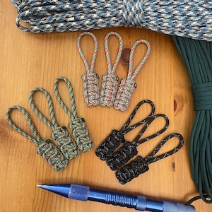 May include: An assortment of paracord zipper pulls in various colors, including black, green, and a mix of red, tan, and white. The pulls are arranged on a wooden surface, with rolls of matching paracord in the background. A blue metal tool is in the foreground.