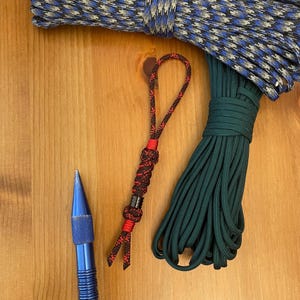 May include: A blue metal tool, a black and red paracord lanyard with a small metal bead, a coil of dark green paracord, and a coil of blue, black, and silver patterned paracord. The items are arranged on a wooden surface for scale reference.