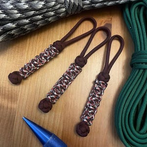 May include: Three brown and white paracord lanyards with a decorative knot at the end. The lanyards are next to a blue metal tool, and rolls of gray and green rope. The lanyards have a loop at the top.