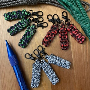 May include: A set of six colorful zipper pulls, each with a different pattern. The zipper pulls are made of paracord and have black metal rings. The zipper pulls are arranged in two rows of three, with the top row showing a green, purple, and black pattern, the middle row showing a red, black, and white pattern, and the bottom row showing a blue, white, and black pattern.