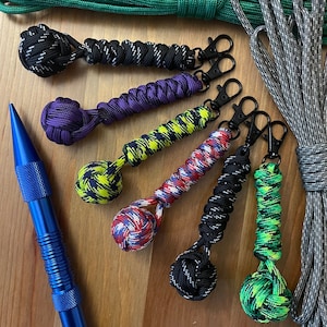 May include: A collection of six colorful keychains with different knot designs. Each keychain features a small ball at the end, with colors including black, green, red, purple, yellow, and white.