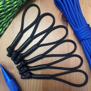 May include: Five black paracord zipper pulls with a loop on each end. The zipper pulls are on a wooden surface.