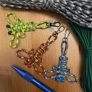 May include: Three colorful paracord keychains in the shape of a dragonfly. The keychains are made with yellow and blue paracord, red and yellow paracord, and blue and gray paracord. Each keychain has a small metal ring and a black metal clasp.