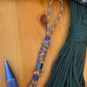 May include: A paracord lanyard featuring a multi-colored rainbow pattern cord with blue accents and a central metallic blue bead. The lanyard is constructed with intricate knots and includes a loop at the top, positioned alongside a blue metal tool.