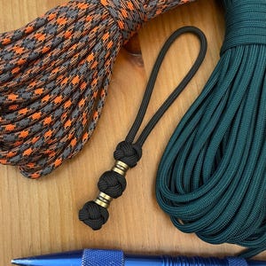 May include: A black paracord lanyard featuring three woven knots and two metallic brass-colored beads, positioned between a coil of orange and gray patterned paracord and a coil of solid dark green paracord, with a blue metal tool at the base.