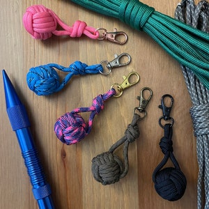 May include: A collection of five colorful keychains with different knot designs. Each keychain features a small, round, knotted ball in a variety of colors, including pink, blue, green, and black. The keychains are made with paracord and have metal clasps.