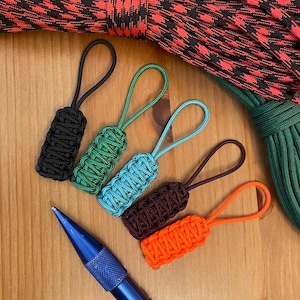 May include: A collection of colorful zipper pulls in black, green, teal, brown, and orange, each with a matching loop. A blue pen is in the foreground. The background includes red and black patterned rope and green rope.