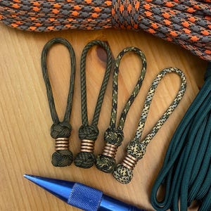 May include: Four paracord zipper pulls in shades of green and camouflage patterns, each with a copper-colored metal accent. A blue tool and two different types of rope are also visible.