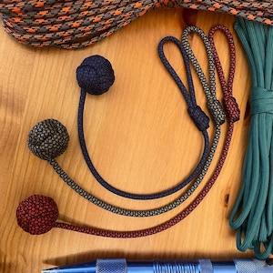 May include: Three paracord lanyards with monkey fist knots in solid navy, black-grey, and red-black patterns. Each features a looped end. Included are a large coil of orange-grey paracord, a coil of teal paracord, and a blue metal tactical pen.