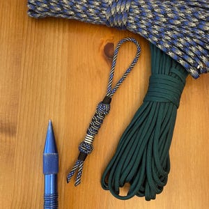 May include: A blue and black paracord lanyard featuring a metallic bead, positioned next to a coiled dark green paracord rope, a coiled blue and black patterned paracord rope, and a blue metal tool with a textured grip handle.