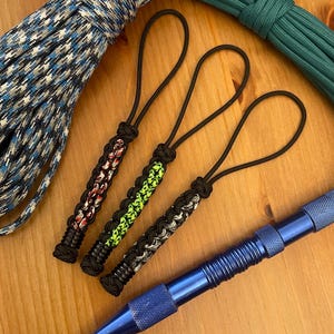 May include: Three paracord keychains with black loops and braided designs in red, green, and gray. A blue metal pen with a pointed tip is also visible, along with rolls of blue and green paracord.