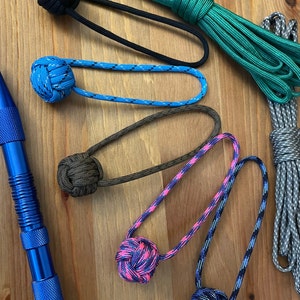 May include: A collection of six colorful knotted rope keychains with different patterns. The keychains are made of paracord and feature a variety of colors, including black, blue, green, brown, pink, and purple.