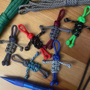 May include: A collection of six colorful paracord keychains, each featuring a unique design. The keychains are made with different colors of paracord and are tied into intricate knots. The keychains are all different shapes and sizes, and some have small beads or charms attached to them.