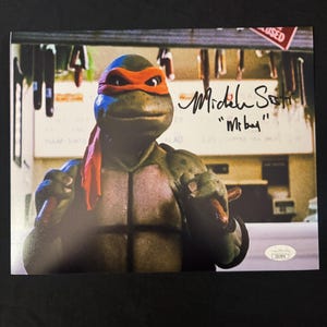 May include: A signed photo of a Teenage Mutant Ninja Turtle character, Michelangelo, wearing a red bandana and a blue mask. The photo is signed "Michelangelo "Mr. Bay" ".