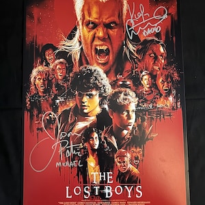 Op de afbeelding: Een rode en zwarte poster voor de film "The Lost Boys" met de hoofdpersonages, waaronder een vampier met tanden, en de titel van de film. De poster is gesigneerd door de acteurs Michael  en Kiefer Sutherland.