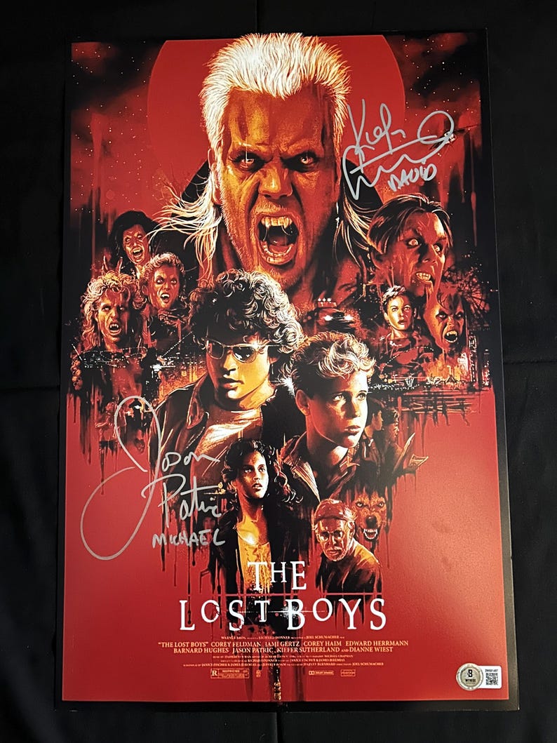 Op de afbeelding: Een rode en zwarte poster voor de film "The Lost Boys" met de hoofdpersonages, waaronder een vampier met tanden, en de titel van de film. De poster is gesigneerd door de acteurs Michael  en Kiefer Sutherland.