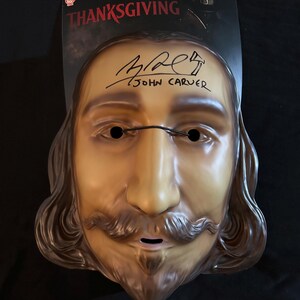 May include: A brown plastic mask of a man with long brown hair, a moustache, and a beard. The mask has black eyes and a black line across the nose. The mask is signed "John Carver" in black marker.