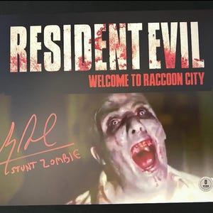 Puede incluir: Un póster firmado para la película Resident Evil, con un zombi con la boca muy abierta. El póster tiene el texto "Resident Evil" y "Bienvenido a Raccoon City". La firma dice "Stunt Zombie".