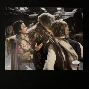 May include: A black and white photograph of three people from the Star Wars film franchise. The photograph is signed by Peter Mayhew, who played Chewbacca. The photograph is authenticated by a Beckett Authentication Services hologram sticker.
