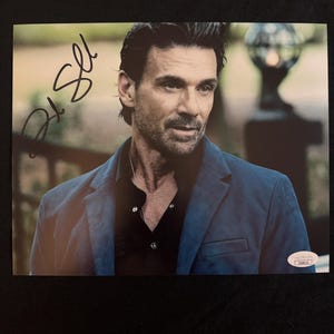 May include: An autographed photograph featuring a man in a dark blue blazer over a black shirt. The photograph has a signature in black ink in the upper left corner. The background is blurred, with a hint of greenery and a decorative lamp post.