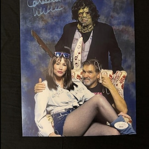 May include: A signed photo of three people, two women and a man, with the man wearing a mask and holding a chainsaw. The woman on the left is wearing a blue denim skirt and a white shirt. The woman on the right is wearing a black dress and fishnet stockings. The man is wearing a black jacket and a white shirt. The photo is signed "Caroline Williams".