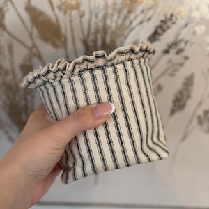 May include: A small, fabric storage basket with a ruffled top. The basket features a cream-colored background with vertical black stripes. The rim of the basket is ruffled, adding a decorative touch. The basket is held in a hand.