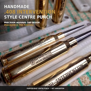 May include: A set of gold-coloured metal centre punches designed in the style of .408 Intervention cartridges. Each tool features engraved text including .408 INTERVENTION, DA MAIN MOUSE, and EXTON INDUSTRIES, with a tapered steel tip.