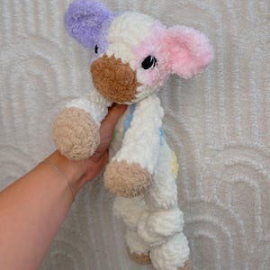 May include: A handmade plush cow toy with a soft, textured appearance. The cow features a color palette of white, lavender, pink, and brown. The ears are lavender and pink, the snout is brown, and the hooves are tan. The toy is designed with black eyes and a friendly expression.