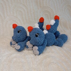 May include: Three blue crochet dinosaur toys with red and white accents. The dinosaurs have gray feet and black eyes. The toys are lying on a white, textured surface.