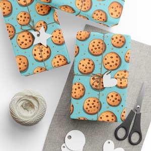 May include: Gift wrap featuring a repeating pattern of chocolate chip cookies on a light blue background. The wrapped gifts are tied with twine and adorned with star and heart-shaped tags. A ball of twine and scissors are also present.