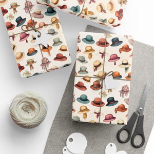 May include: Gift-wrapped boxes with a repeating pattern of watercolor hats in various styles and colors, including red, blue, and tan. The boxes are tied with twine and adorned with heart-shaped tags. A ball of twine, scissors, and blank tags are also present.