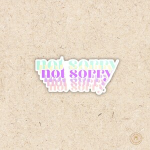 May include: A white sticker with the words "not sorry" in a layered design. The text is in pastel colors of green, purple, and yellow. The sticker is on a light brown textured background.