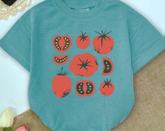 Peut inclure: Un t-shirt à manches courtes et col rond de couleur bleu sarcelle atténué. L'avant du vêtement présente une impression graphique de diverses tomates rouges, incluant des tomates entières, des moitiés et des tranches, disposées en motif.