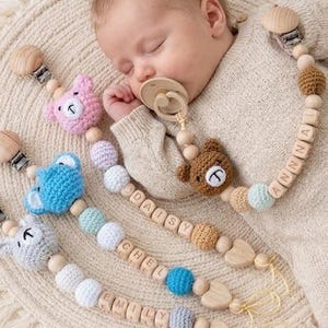 May include: Four personalized pacifier clips featuring crocheted bear heads in pink, blue, and gray, combined with wooden beads, lettered blocks spelling names, and metal clips. One clip is attached to a pacifier in a sleeping infant's mouth.
