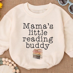 May include: Cream-colored baby romper with the text "Mama's little reading buddy" in black. A stack of books is printed below the text. The romper has long sleeves and a round neckline. A pair of light blue sunglasses and a wooden beaded necklace are also in the image.